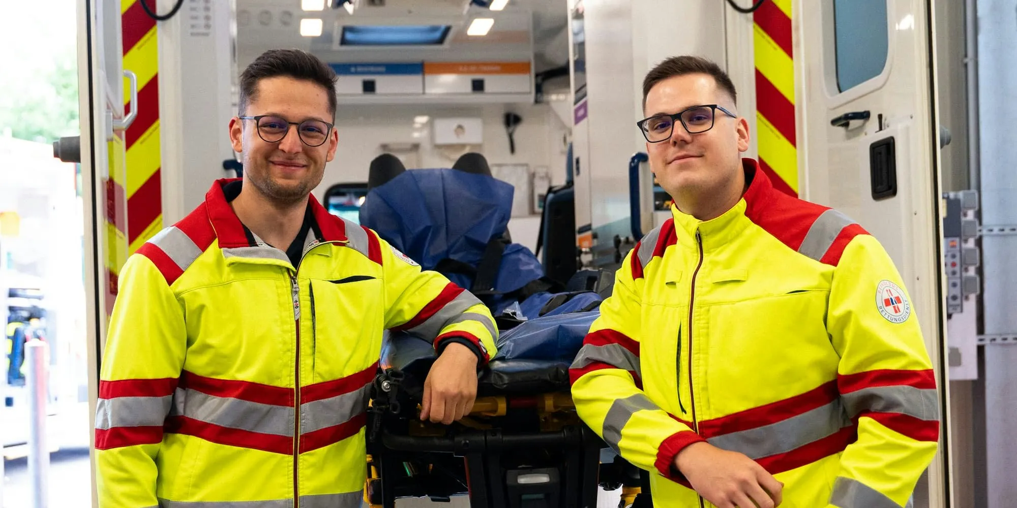 Zwei Sanitäter in gelb-roter Dienstkleidung stehen vor einem Rettungswagen mit offener Hecktür. Beide lehnen sich an die ausgefahrene Trage an und lächeln in die Kamera